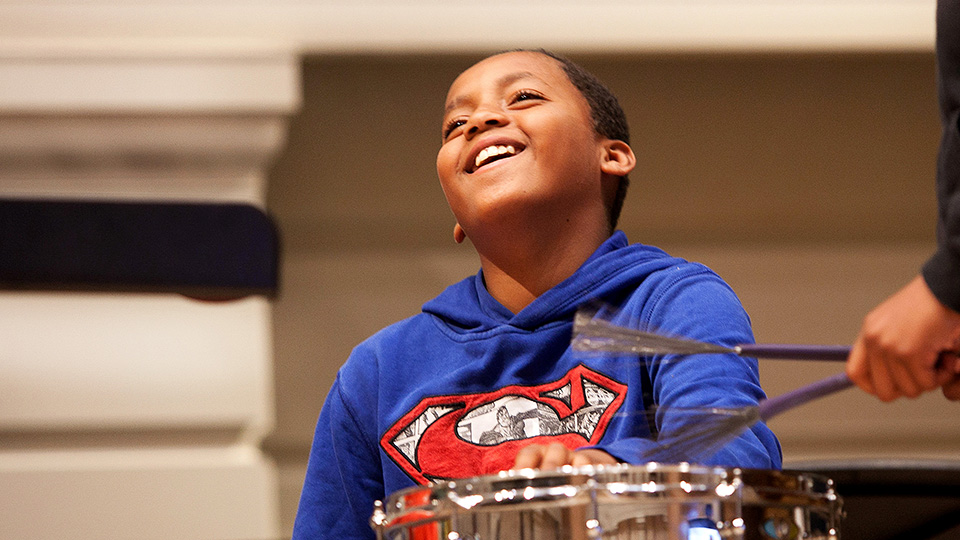 Child at a Sparks event laughing whilst playing the percussion, with someone else playing percussion in the foreground